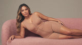 Woman wearing a Stage 2 post-surgical faja with full torso compression on a pink boucle sofa, showing coverage from underbust to hip after lipo 360
